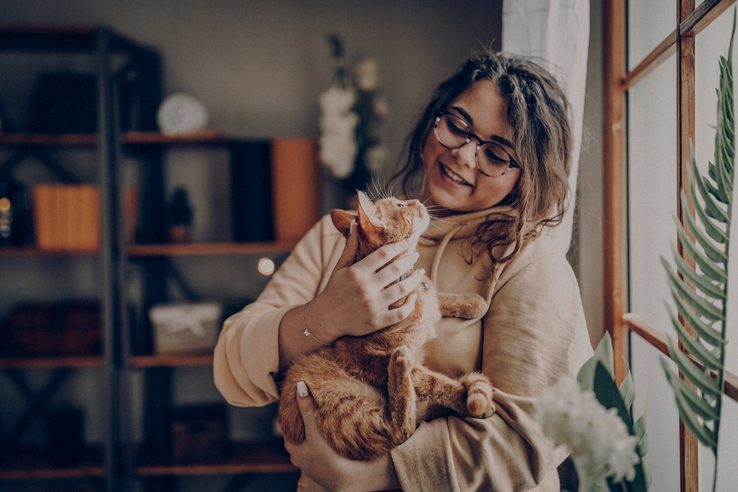 Smiling woman in glasses cuddling an orange cat indoors